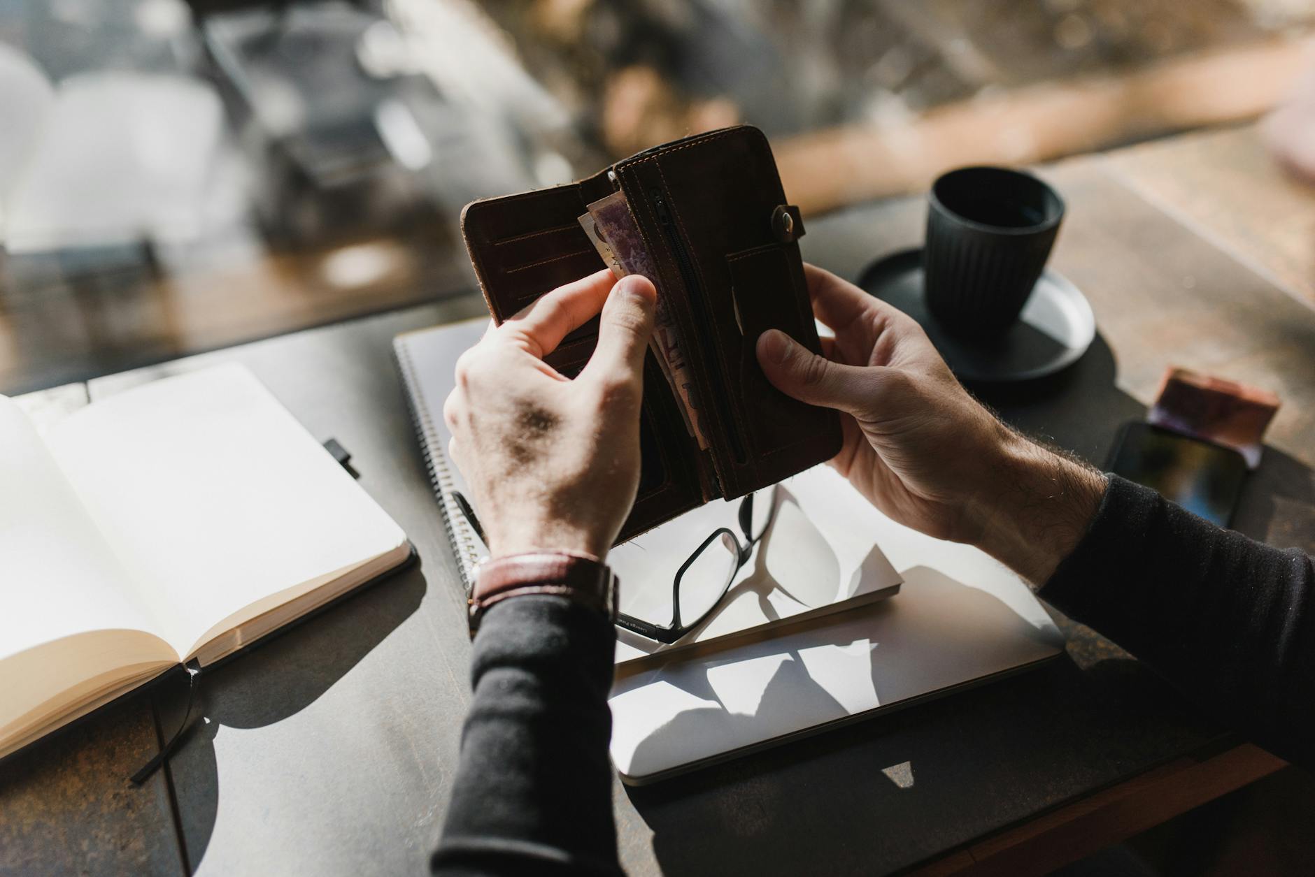 Portfel z gotówką na biurku obok okularów, notesu i filiżanki kawy