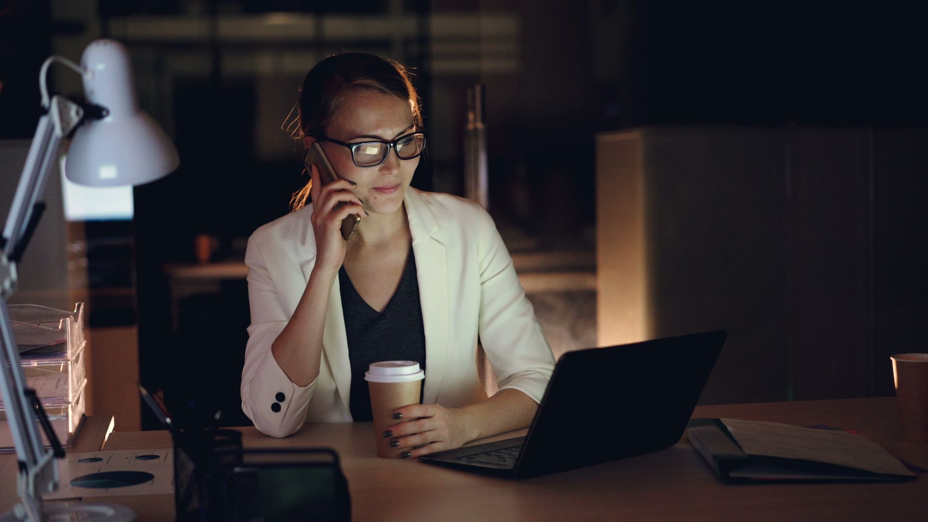 Kobieta w okularach pracuje nocą przy laptopie z kawą i telefonem